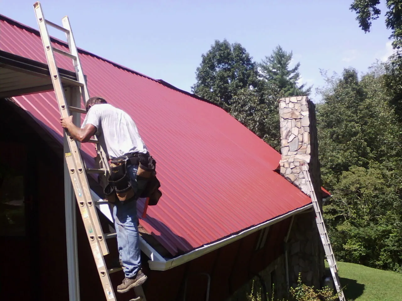 Roofer on a ladder beside a red metal roof and stone chimney — Soffit & Fascia Repair by Roofing Brothers in Mcadoo